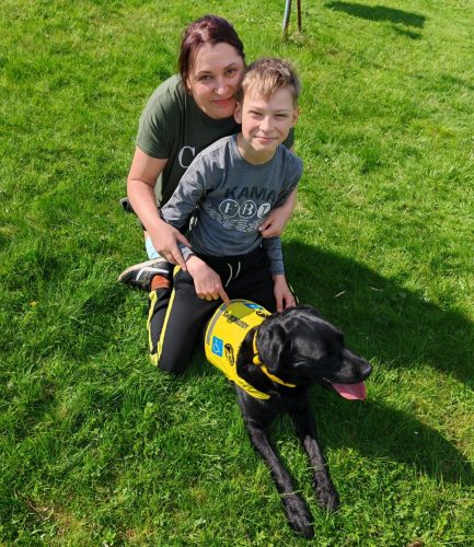 Chlapec sedící na trávě vedle své matky a asistenčního psa