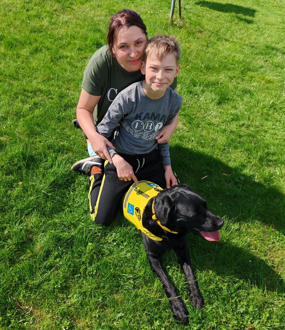 Chlapec sedící na trávě vedle své matky a asistenčního psa