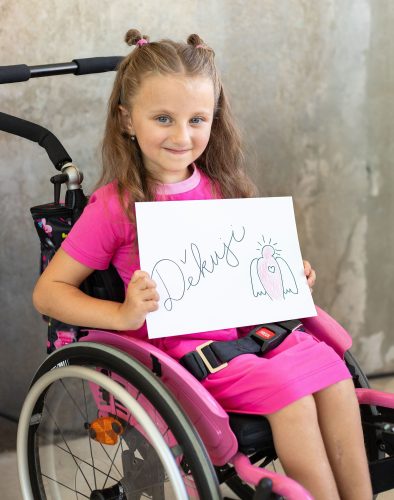 2025_zakulisi-18 Nikolka sedící na vozíčku a držící nápis Děkuji s nakresleným logem Dobrého anděla